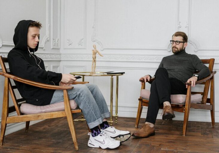 A concerned teenage boy in a hoodie sits with a therapist during a session.