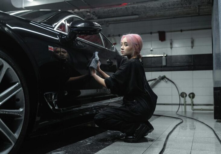 A young female worker polishing a black luxury car in an indoor garage setting.
