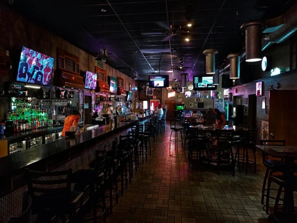A dimly lit modern bar interior featuring a long counter and multiple screens, creating a cozy urban atmosphere.